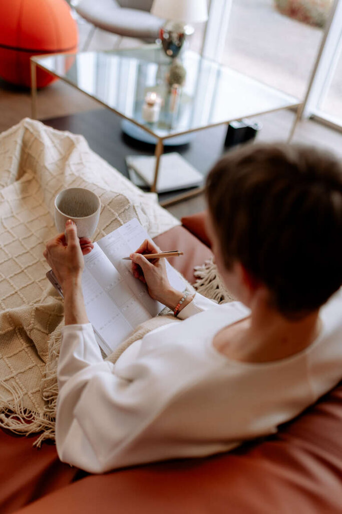 Frau schreibt am Fenster in ihren Terminkalender, eine Tasse in der Hand.