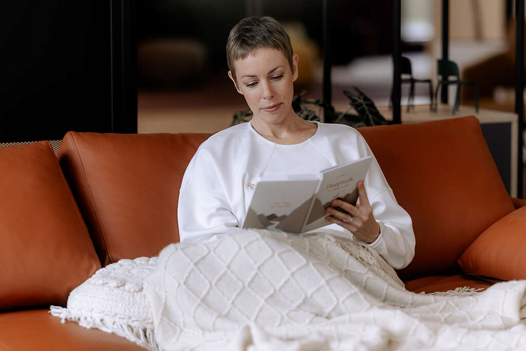 Frau mit kurzen Haaren liest in eine Decke gehüllt auf einem orangefarbenen Sofa.