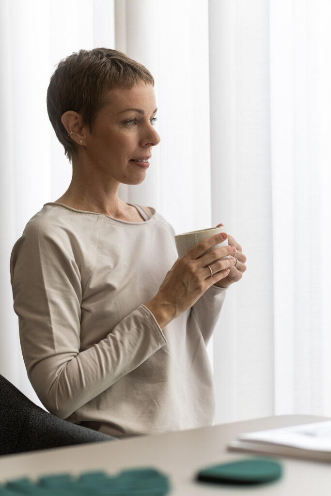 Eine Frau mit kurzen Haaren sitzt mit einer Tasse in der Hand am Fenster und wirkt nachdenklich. Sie trägt ein helles Langarmshirt, und hinter ihr fällt sanftes Licht durch die durchsichtigen Vorhänge – ihr nachdenklicher Blick lässt vermuten, dass sie vielleicht über hormonelle Beschwerden in Göttingen und die damit verbundenen Herausforderungen nachdenkt.